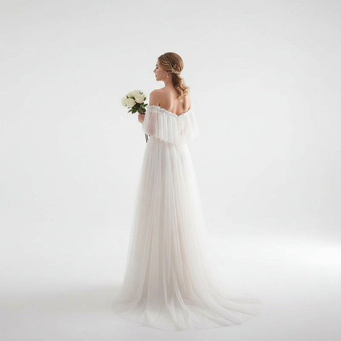 Woman in a white wedding dress holding flowers in a bright room with sheer curtains.