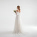 Woman in a white wedding dress holding flowers in a bright room with sheer curtains.