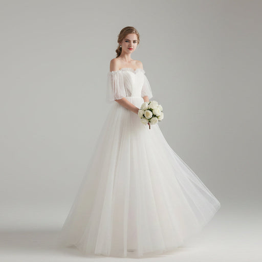 Woman in a white wedding dress holding flowers in a bright room with white curtains.