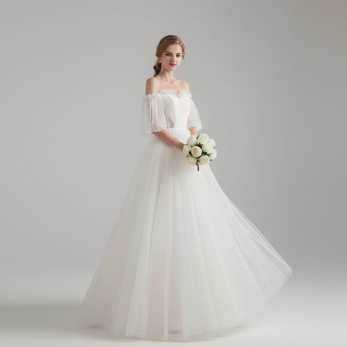 Woman in a white wedding dress holding flowers in a bright room with white curtains.