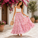 A woman stands outdoors in a sunlit courtyard wearing a white spaghetti-strap crop top and a long pink floral maxi skirt, holding out the skirt to show its flow. She carries a woven straw tote and wears beige sandals.